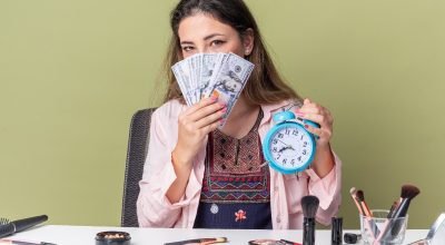 pleased-young-brunette-girl-sitting-table-with-makeup-tools-holding-money-alarm-clock (1)